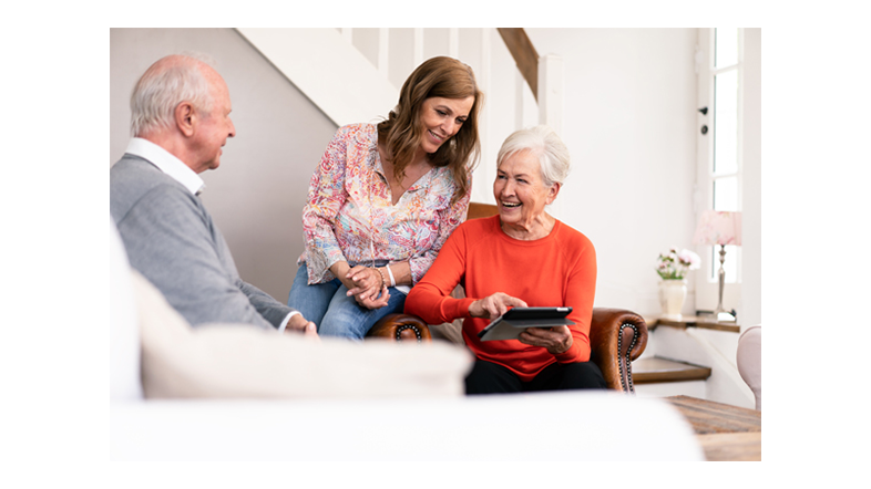Ältere Frau zeigt lächelnd etwas auf einem Tablet, während eine jüngere Frau daneben steht und hilft