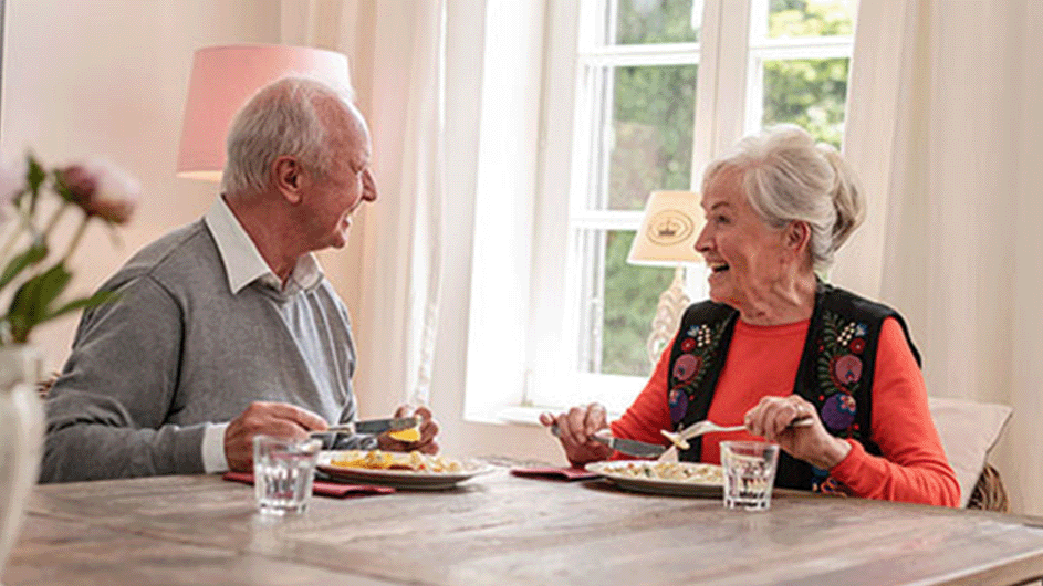 Älteres Paar sitzt am Tisch beim Essen und lacht sich an